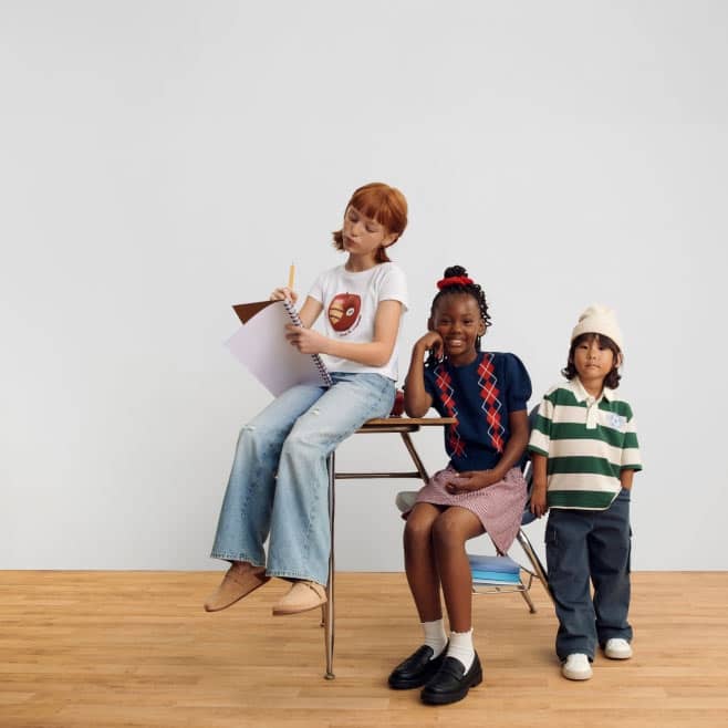 Three children wearing a graphic tee with wide leg jeans, an argyle sweater with skirt and a striped polo with pants and a beanie.