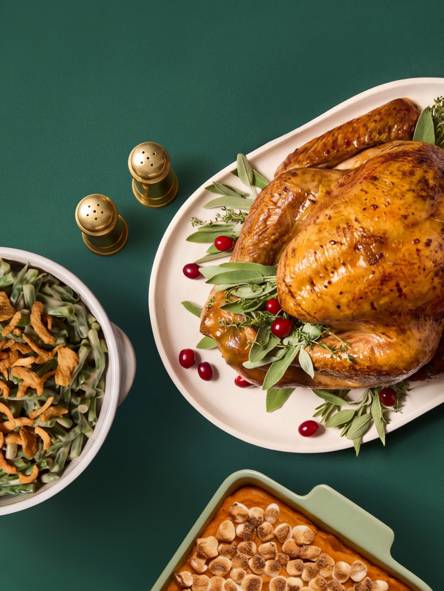 Roast turkey with herbs and cranberries on a platter with green bean casserole, sweet potatoes with marshmallows and gold salt and pepper shakers.