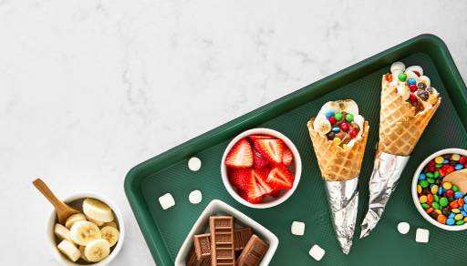 Two cones full of mini marshmallows and candy next to sliced strawberries and bananas, chocolate pieces, and candy-coated chocolates.