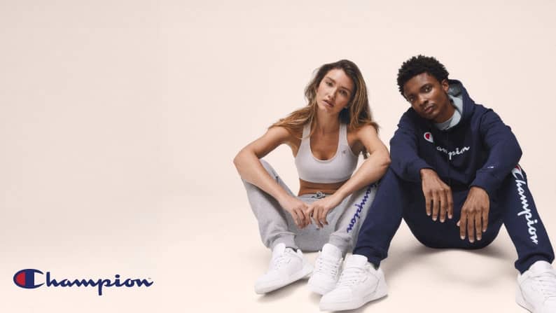 Champion logo in the bottom left corner. A woman and a man wearing Champion looks are sitting on the ground.