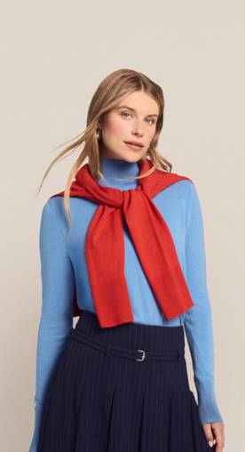A woman wearing a blue sweater with a red sweater tied around her shoulder is pictured on a beige backdrop.