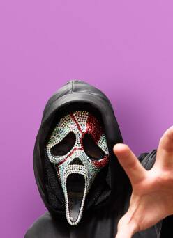 Person wearing a black hood and a scary screamer mask featuring white and red crystals. They are reaching out with one hand.