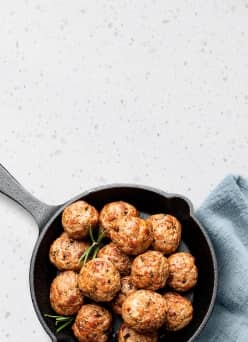 A skillet of meatballs and herbs on a light blue kitchen towel.