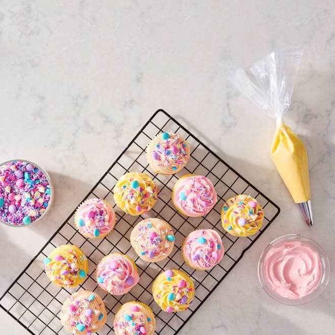 A rack of cupcakes with yellow and pink frosting and sprinkles next to a bag of yellow frosting, a bowl of pink frosting and a bowl of sprinkles.