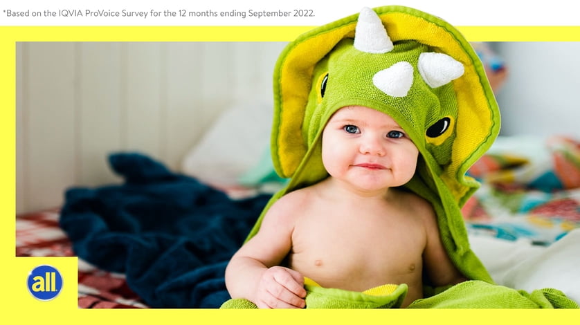 A baby sits on a bed while swaddled in a dinosaur blanket.