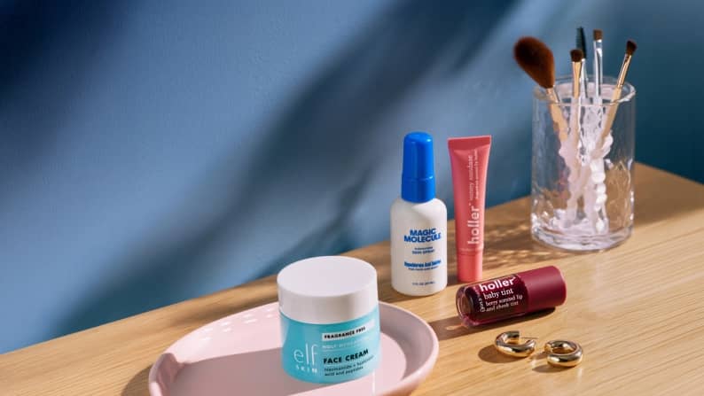 A collection of beauty products, makeup brushes and earrings is on top of a wooden desk in front of a blue wall.
