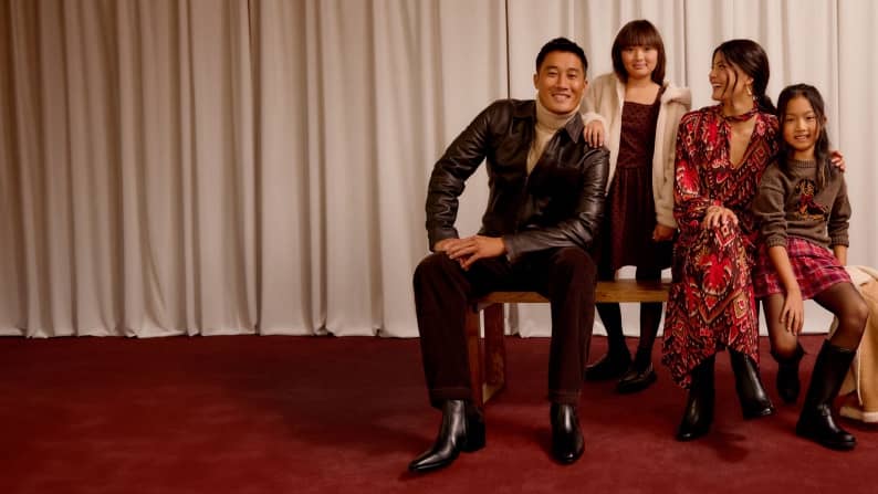 Family of four in fall outfits sits and stands on a wooden bench in front of a curtain backdrop, smiling together.