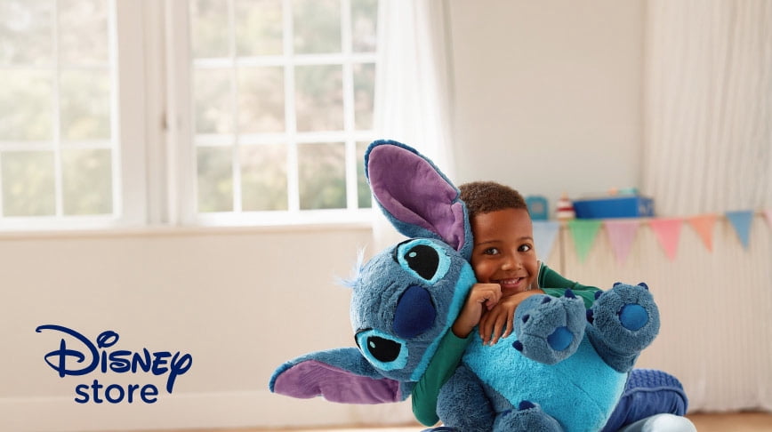 Disney store. A smiling child hugs a large blue Stitch plush toy in a bright room with colorful pennant decorations.