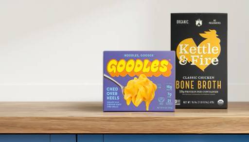 Goodles ched over heels macaroni and cheese next to Kettle and Fire classic organic chicken bone broth on a wooden counter.