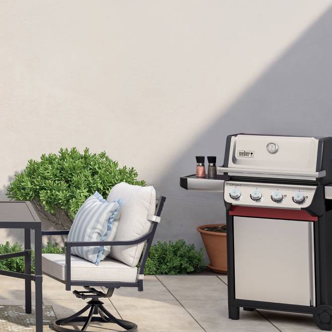 A grill situated next to green plants as well as black and white outdoor furniture, atop a white floor.