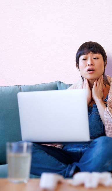 A woman touching her neck, wrapped in a blanket accessing virtual care from her laptop.