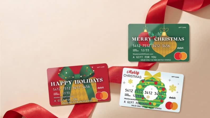 Mastercard debit gift cards with Happy Holidays and Merry Christmas designs arranged beside a curling red ribbon.