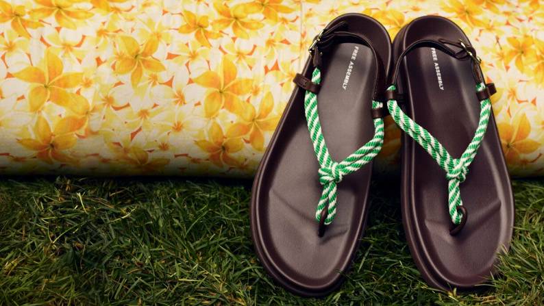 Brown thong sandals with green-and-white braided straps and toe loop, set on grass against a yellow floral backdrop.