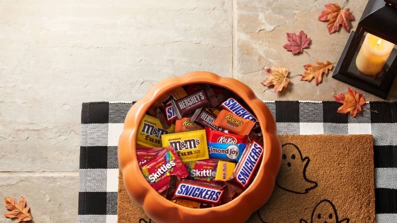 Pumpkin bowl filled with assorted Halloween candy on a checkered cloth and ghost doormat beside a lantern and autumn leaves.