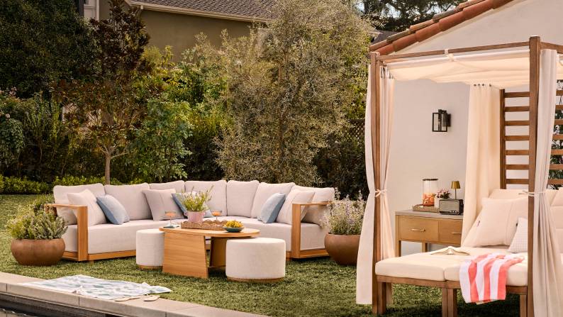 An outdoor sectional and a cabana sit on a lawn near a pool. The scene is styled with blue pillows, a wood coffee table and plants.