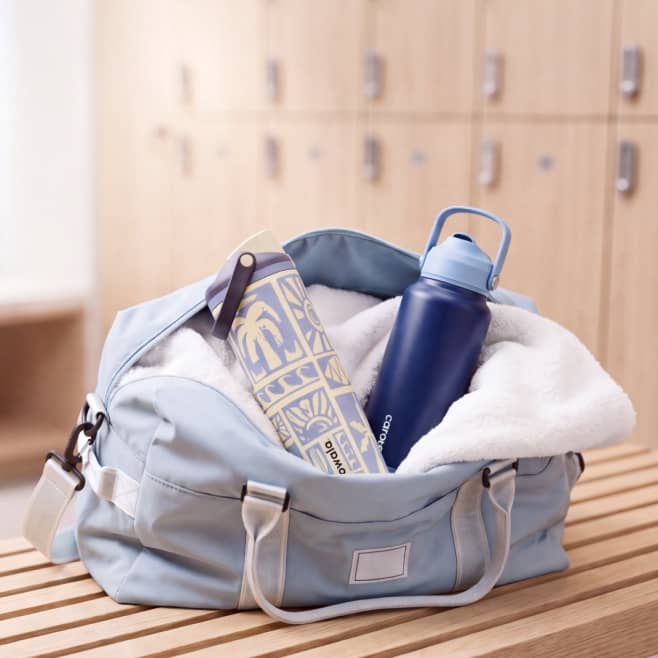 Light blue gym bag on bench holding patterned water bottle, white towel and blue water bottle.