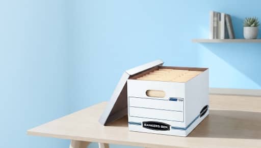 A cardboard filing box on a wood desk, against a blue wall.