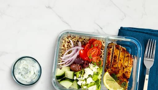 Grilled chicken, brown rice and fresh veggies are displayed in a glass storage container. A cup of yogurt dipping sauce is also shown.