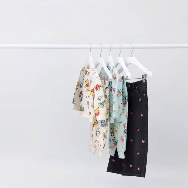 Children shirts with cartoon prints and one pair of dark jeans with pink hearts hang on white hangers on a simple clothing rack.