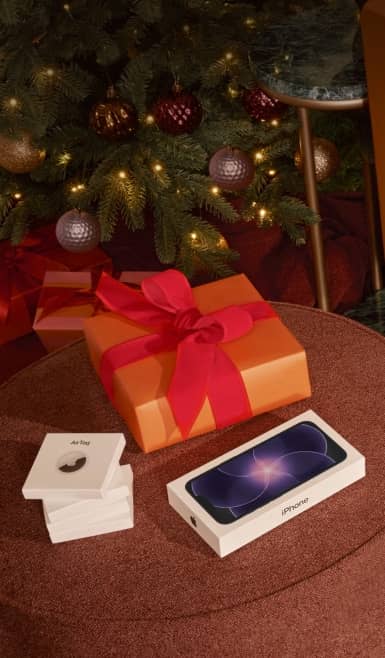 Apple iPhone and AirPods boxes on a round table with a large orange gift box in front of a lit Christmas tree.
