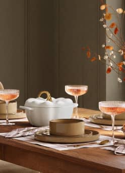 Warm fall tabletop with pumpkin shaped serving dish, layered plates and napkins, and coupe glasses filled with a rose colored drink.
