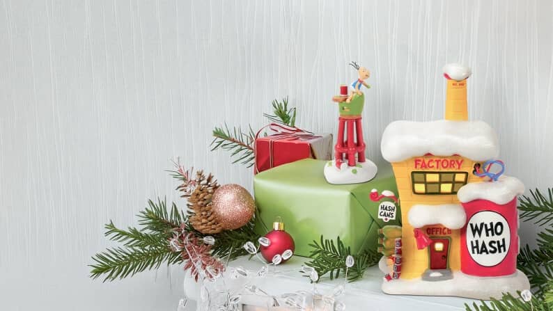 Christmas village decor with Factory building, Who Hash container, wrapped gifts, ornaments and evergreen branches on a snowy base.