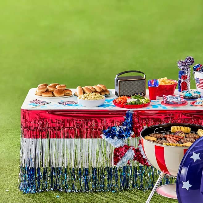 Memorial Day BBQ with red, white and blue foil fringe table decoration, hamburgers, hot dogs, napkins, decor and Americana themed grill.