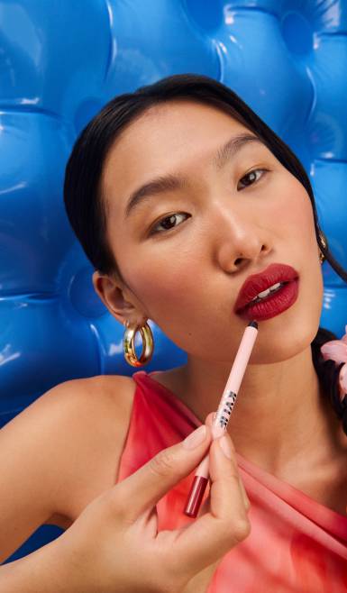 Woman with a matte red lip applies a lip liner against a bright blue background.