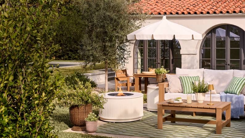 A fluted white fire pit and wooden patio furniture with green pillows sit on a patterned rug beneath a white umbrella in a Mediterranean-style yard.