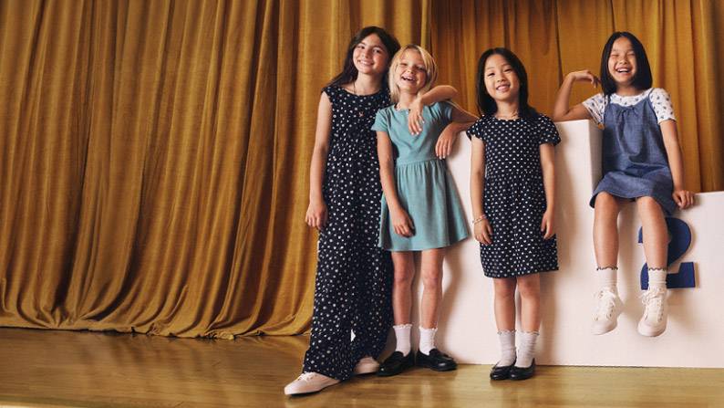 Four children in dresses and a jumper smile together on a stage, with one sitting on a white platform marked with the number two.