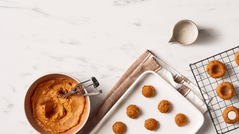 A bowl full of cookie dough with a measuring scoop next to a tray of unbaked cookies, and a cooling rack of thumbprint cookies.