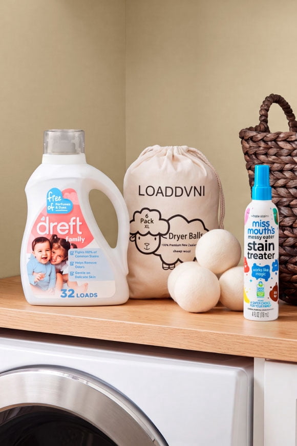 Dreft laundry detergent, Loaddvni dryer balls and Miss Mouths Messy Eater stain treater on a wooden shelf above a washing machine.