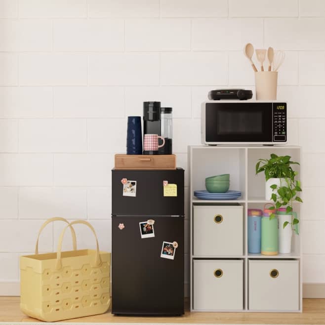 A mini fridge sits by a white shelf holding plates, bins, and water bottles. A microwave and coffee maker are on top.