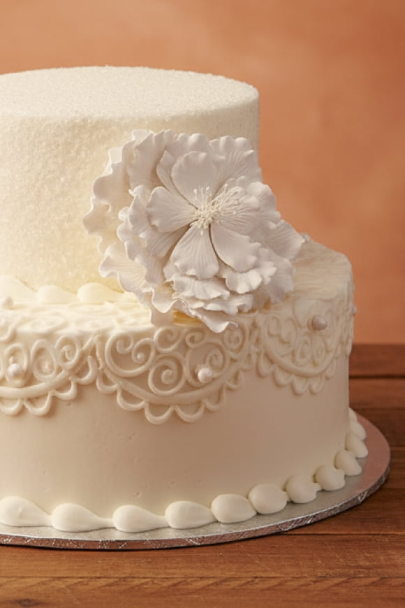 A two-tiered white cake is shown with intricate white piping and a white flower.