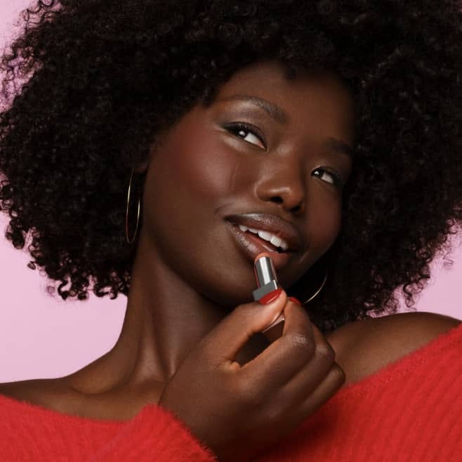 A close-up model shot of a woman wearing a red top. She’s looking away and applying red sheer lipstick.