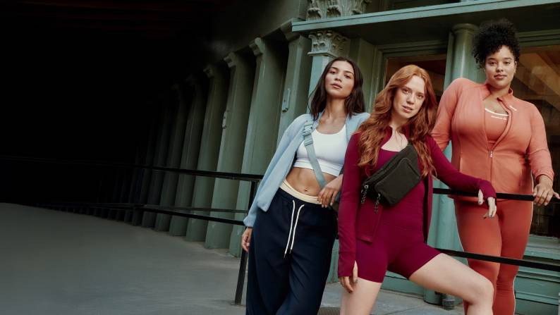 Three young women shown in front of a building wearing workout tanks, sports bras, zip-up jackets and leggings from Athletic Works, Avia, Reebok.