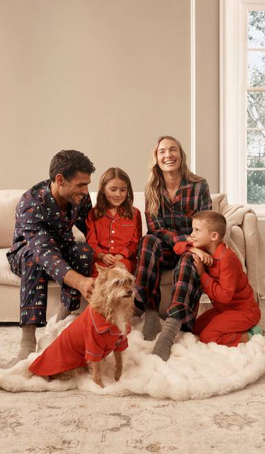 Family of two adults and two children in festive pajamas on a sofa with a small dog in a red outfit on a fluffy rug.