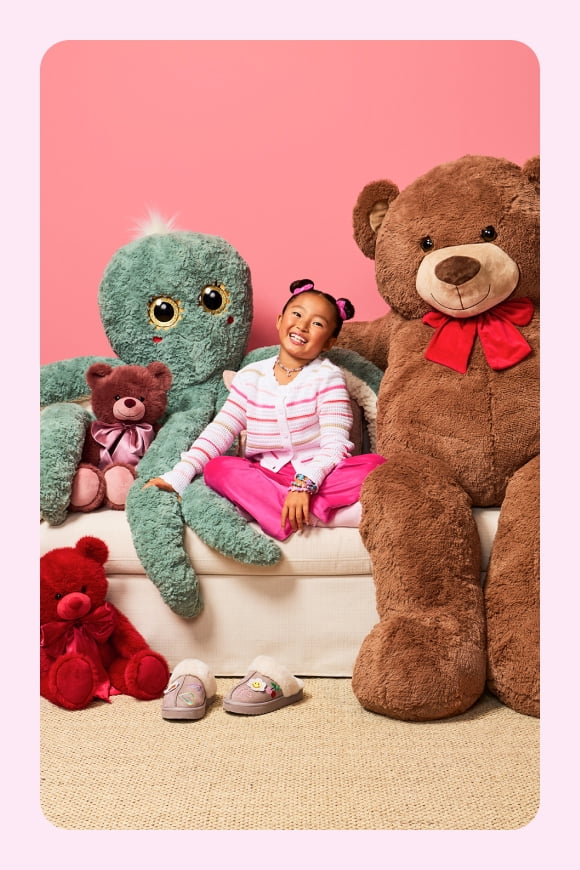 A kid sits between huge life-sized plushies of an octopus and a bear. Smaller plush bears are arranged around her.
