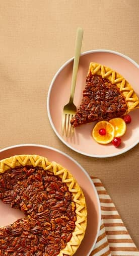 A pecan pie with a slice missing is shown next to a slice of pecan pie on a pink plate garnished with orange slices and cranberries.