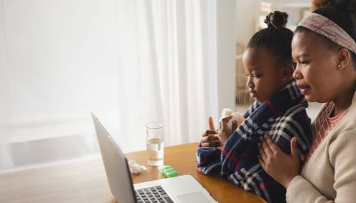 A mother sits beside her child, comforting them while they look at a laptop during a telehealth visit.