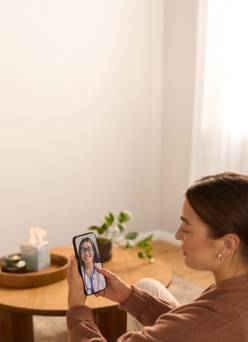 An adult sits on a sofa holding a smartphone showing a doctor with a stethoscope on a video call in a living room.