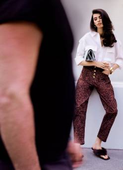 Woman in a white blouse tucked into burgundy printed jeans with fuzzy sandals and a black clutch.