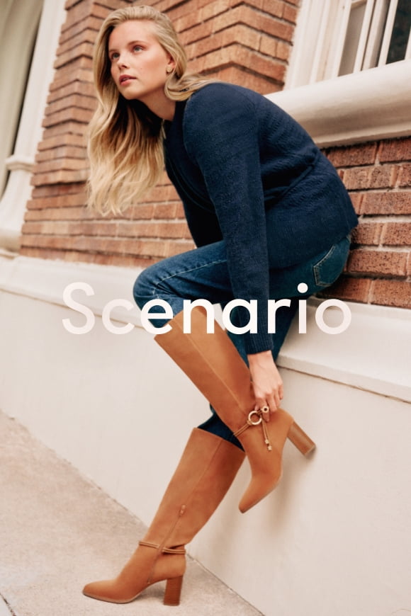 Woman wearing tan knee high boots with ankle wrap details, blue jeans and a navy sweater.
