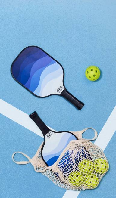 Rackets and balls spill out of a netted bag and onto a pickleball court.