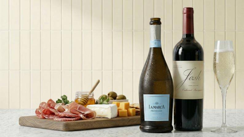 A stocked charcuterie board next to a bottle of LaMarca prosecco, a bottle of Josh cabernet sauvignon, and a glass of prosecco.