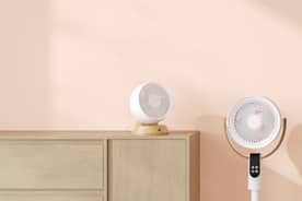 Two white fans, one on the floor and another atop a brown wood table against a pink wall.