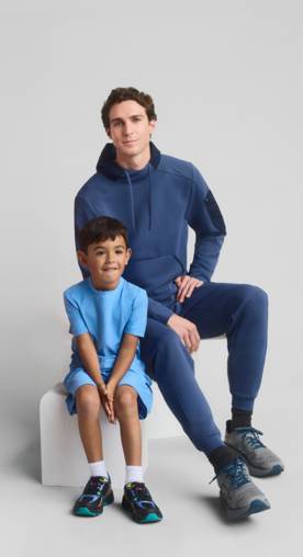 A father and son are shown sitting on a white block against a white, grey background wearing a hoodie, tee, shorts and joggers.