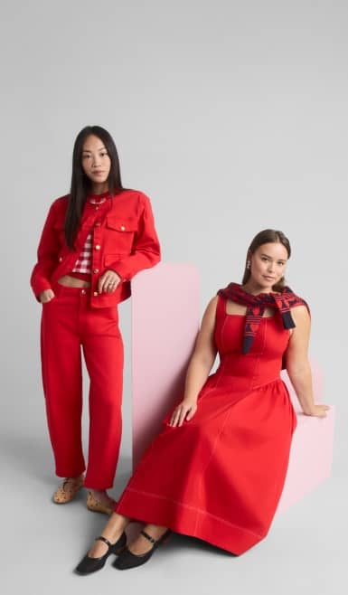 A woman wearing a red jacket and pants is standing next to a woman wearing a red dress who is sitting in a white block.