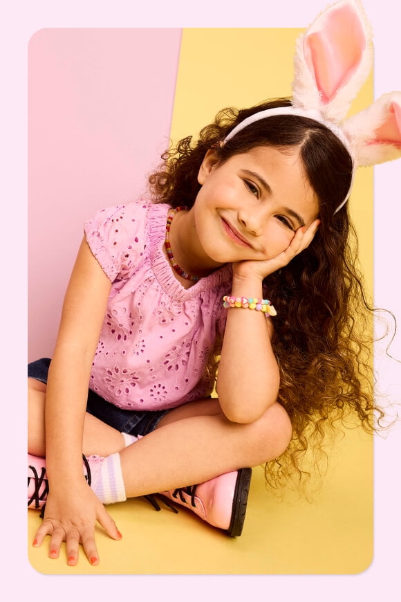 A child with a pink shirt and bunny ears sits against a pink and yellow background
