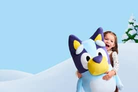Girl shown hugging large Bluey plush toy against a snowy background. Bluey logo also shown.